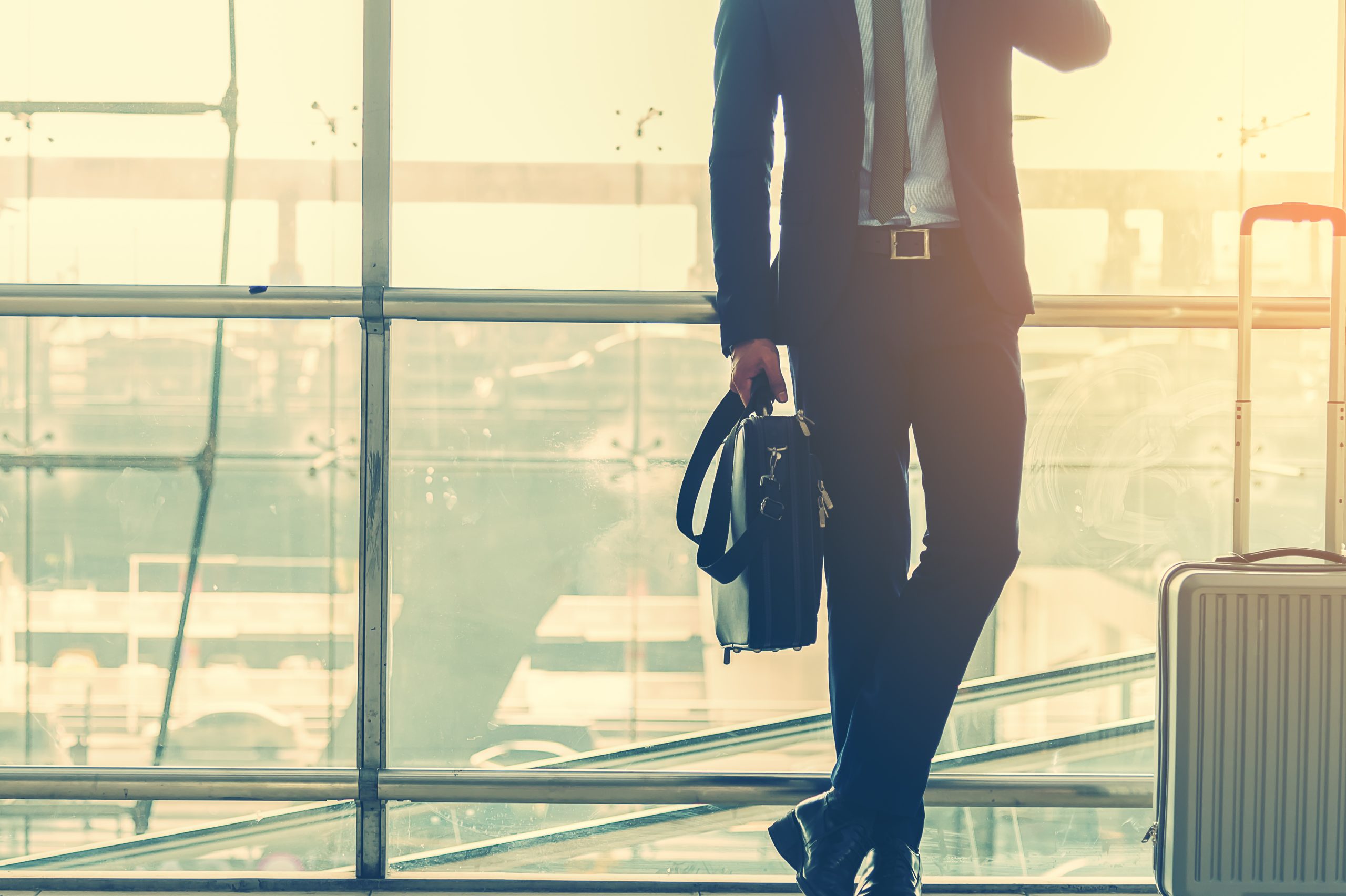 Businessman with a baggage talking via mobile phone at the airport. Businessman wearing suit jacket indoors. A man standing with a baggage using a smart phone in airport departure terminal. Business